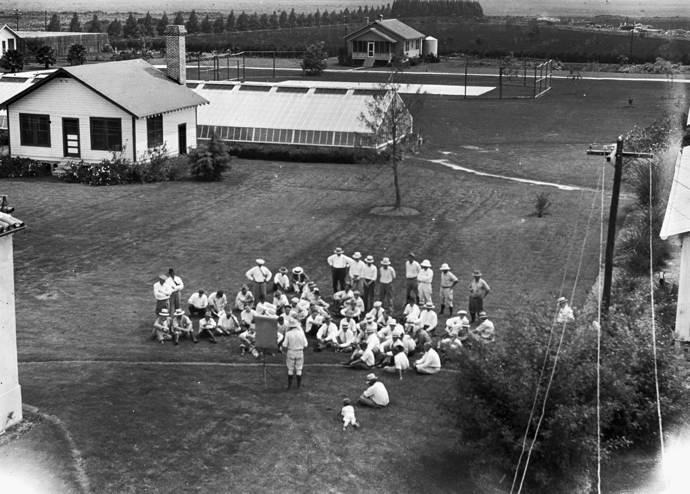 Overhead view of about fifty bean growers in hats on the lawn behind the Everglades Experiment Station lab, facing a man at an easel, with a young boy crawling on the grass nearby